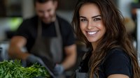 Young Caucasian woman with bright smile holding fresh herbs in modern restaurant kitchen while male chef works in background. Professional culinary setting.