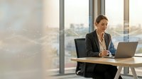 Professional businesswoman working on laptop in modern office with city view through large windows during golden hour lighting.
