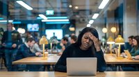 Stressed Asian businesswoman holding head in hands while working on laptop in modern office coworking space during evening hours.