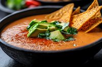 Creamy orange soup garnished with fresh avocado slices, cilantro and tortilla chips in black bowl. Traditional Mexican cuisine with paprika powder on top.
