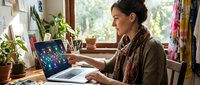 Woman with dark hair and olive complexion working on laptop at wooden desk near window with plants and natural light, suitable for remote work, freelance, productivity, and home office concepts.