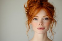 Young woman with natural red hair in messy updo style, blue eyes, and freckles smiling warmly against light background. Soft beauty portrait with minimal makeup.
