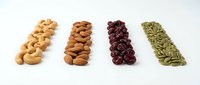 Four rows of healthy nuts and seeds including cashews almonds dried cranberries and pumpkin seeds arranged on white background for snacking and cooking.