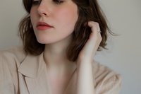 Young Caucasian woman with brown hair and red lipstick touching her hair, wearing beige blazer against neutral background, soft natural lighting creates gentle mood.