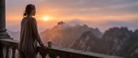 Woman in brown cloak standing on balcony overlooking misty mountain landscape at sunrise, contemplative travel and adventure scene.