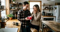 Young couple sharing romantic moment in modern kitchen as man surprises woman with red gift box during morning coffee time together.