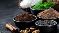 Fresh cocoa powder in black ceramic bowls with mint leaves, cinnamon sticks, walnuts and black sesame seeds on dark stone background, selective focus.