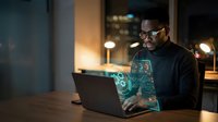 African American businessman working late at night on laptop with holographic digital interface technology in modern office environment.