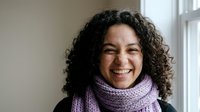 Happy young Hispanic woman with curly hair wearing purple knitted scarf smiling warmly by window in cozy indoor setting for lifestyle and portrait concepts.