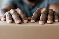 Close-up view of African American male hands resting on cardboard box surface, showing texture and detail of fingers against brown packaging material.