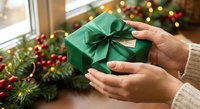 Hands holding green wrapped Christmas gift with bow near festive garland and twinkling lights for holiday celebration and seasonal giving traditions.