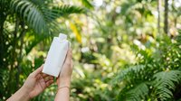 White cosmetic bottle held in hand against lush green foliage background. Suitable for skincare, sunscreen, lotion, and beauty product packaging mockups.