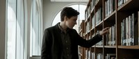 Young Caucasian male student browsing books on wooden library shelves near window, academic research and education concept for university materials.