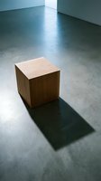 Wooden cube on polished concrete floor in minimalist gallery space with white walls and natural light casting sharp shadow.