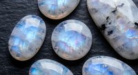 Polished rainbow moonstone gemstones with blue flash adularescence on dark slate background for jewelry making and crystal healing practices.