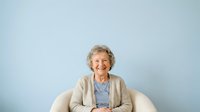 Senior woman with curly gray hair and fair complexion, wearing casual beige cardigan over blue striped shirt, seated in white chair against light blue background for portrait use.