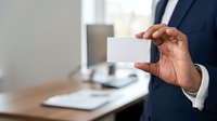 Businessman in dark suit holding blank white business card at office desk with computer and documents in background.