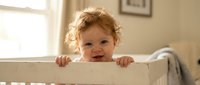 Adorable toddler with curly blonde hair standing in white wooden crib, smiling at camera in bright nursery room with natural lighting and cozy atmosphere.