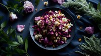 Dried rose petals and chamomile flowers in black ceramic bowl on dark textile background with fresh roses and greenery, moody floral composition.