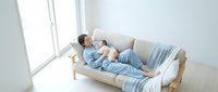 Young Asian woman in blue pajamas relaxing on modern beige sofa by large window in bright minimalist living room for lifestyle and comfort concepts.