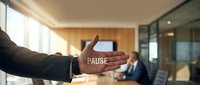 Professional businessman showing pause hand gesture in modern office meeting room with colleagues discussing in background for workplace communication.