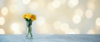Yellow flowers in glass vase on white surface with warm bokeh lights background creating cozy home decor atmosphere for interior design projects.