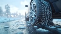 Close-up view of winter tire with deep tread pattern on snowy road surface during cold weather, with bokeh lights and vehicle in soft focus background.