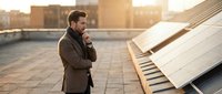Thoughtful male professional in brown jacket standing on rooftop with solar panels during golden hour sunset, contemplating renewable energy solutions for sustainable business development.