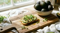 Avocado toast on wooden cutting board with fresh avocados in bowl, natural kitchen setting with window light, food preparation and healthy eating concept.