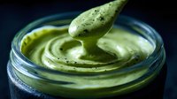 Creamy avocado sauce dripping from spoon into glass jar, rich green texture with black pepper specks against dark background.