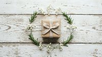Kraft paper gift box with natural bow surrounded by rosemary sprigs and white flowers on rustic white wooden background for holiday celebrations.