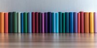 Row of colorful hardcover books standing in line on wooden surface against light wall, creating rainbow spectrum pattern with soft reflection below.