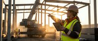 Construction worker in safety vest using tablet at building site with crane and steel framework during golden hour sunset for project management and planning.