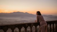 Woman in casual beige sweater leaning on stone balustrade overlooking misty mountain valley at golden hour sunrise or sunset, contemplative mood, travel and nature lifestyle.