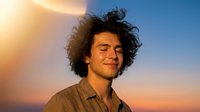 Young Hispanic man with curly hair smiling at sunset with warm golden light and blue sky background for lifestyle and portrait photography.