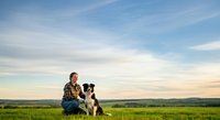 Young adult male farmer in plaid shirt sitting with border collie dog in green pasture under blue sky. Pet companionship and rural lifestyle.