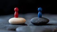 Red and blue wooden figures standing on contrasting light and dark stones against black background, symbolizing balance, opposition, and harmony in business concepts.