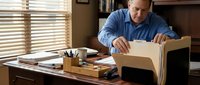 Middle-aged Caucasian businessman packing office belongings into cardboard box at wooden desk. Corporate downsizing, job transition, retirement concept.