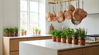 Modern kitchen interior with copper pots hanging from rack and fresh herbs in terracotta pots on white countertop for culinary design projects.