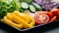 Yellow bell peppers and sliced tomatoes arranged on black plate with fresh vegetables including purple cabbage and cucumber in background for healthy eating and nutrition concepts.