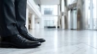 Businessman legs in polished black leather shoes and dark suit trousers standing on marble floor in modern corporate office lobby.