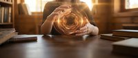 Hands holding glowing orb of light on wooden desk with books and warm interior lighting, representing innovation, creativity, and business ideas.