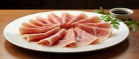 Thinly sliced prosciutto ham arranged on white plate with fresh herbs and balsamic sauce for gourmet appetizer and charcuterie board presentation.