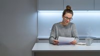 Young professional woman with glasses reading documents in modern kitchen with white countertops and LED lighting for home office work.