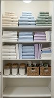 Organized linen closet with neatly stacked towels in cream, blue and green, folded sheets, woven storage baskets and glass containers on white shelving for home organization and interior design.