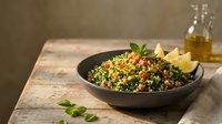Fresh tabbouleh salad with parsley, tomatoes and bulgur wheat served in dark bowl on rustic wooden table with olive oil and lemon wedges.
