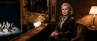 Elegant senior Caucasian woman in luxury theater box wearing formal black attire with sophisticated styling during evening performance.