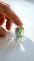 Hand holding colorful glass marble with swirled orange, blue, and yellow patterns, casting vibrant light reflections on white surface for play, collection, or nostalgic themes.