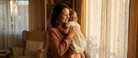 Young African American mother tenderly holding and embracing her infant baby in warm sunlit living room, expressing love and maternal bond at home.