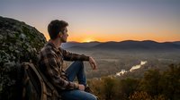 Man in casual plaid shirt and backpack sitting on rocky mountain overlook at sunset, gazing at valley landscape with river and distant mountains, contemplative outdoor adventure scene.